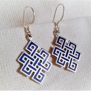 Puede incluir: Un par de pendientes de plata con un diseño de esmalte azul. El diseño es un patrón de nudo tradicional, a menudo utilizado en el arte tibetano y budista.