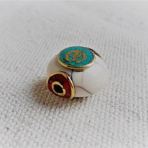 May include: White bead with a turquoise and gold Om symbol and a red and gold accent. The bead has a hole for stringing.