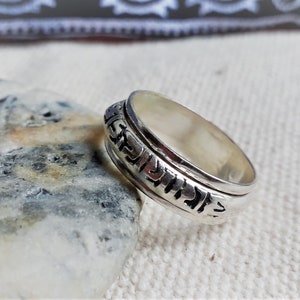 May include: A silver spinning ring with an inscription in a foreign language. The ring is on a gray and white stone.