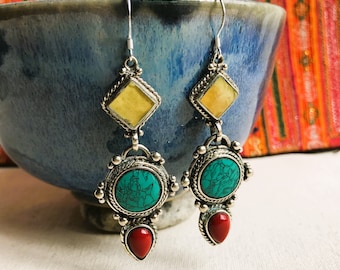 May include: A pair of silver dangle earrings featuring turquoise and red stones. The earrings have a square-shaped stone at the top, followed by a round turquoise stone, and a teardrop-shaped red stone at the bottom.