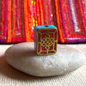 Peut inclure: Une perle rectangulaire avec un dessus et des côtés turquoise, un centre rouge et un motif géométrique doré. La perle repose sur une pierre grise lisse, avec un textile coloré en arrière-plan.