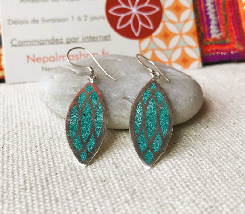 May include: A pair of silver-toned earrings with a leaf-like shape. Each earring features a teal glitter-filled design within a silver frame. The earrings are displayed on a white surface.