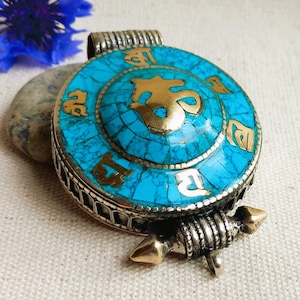 Può includere: Un ciondolo rotondo in turchese e oro con una catena in argento e oro. Il ciondolo presenta un'intarsio in turchese e un'iscrizione dorata in scrittura tibetana.