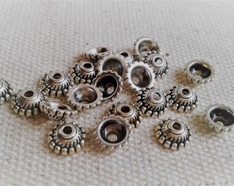 May include: A collection of silver-colored metal bead caps with a detailed, textured design. Each cap has a conical shape with a decorative edge and a small hole at the top. The beads are scattered on a white, textured surface.
