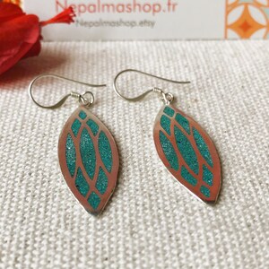 May include: Silver-tone dangle earrings with a leaf-shaped design. The earrings feature a copper-colored outline with a turquoise-colored, textured inlay. The earrings have a simple hook closure and are displayed on a textured, off-white surface.