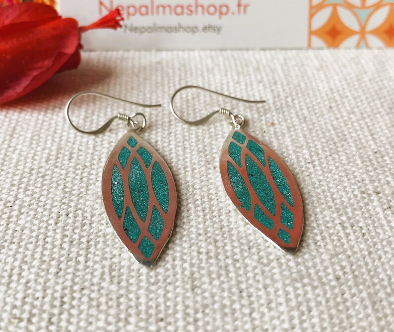 May include: Silver-tone dangle earrings with a leaf-shaped design. The earrings feature a copper-colored outline with a turquoise-colored, textured inlay. The earrings have a simple hook closure and are displayed on a textured, off-white surface.
