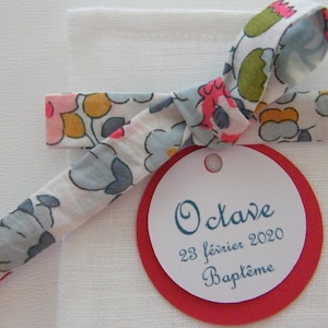 Puede incluir: Una bolsa de regalo de tela blanca atada con una cinta floral y una etiqueta redonda roja con el texto "Octave 23 février 2020 Baptême".