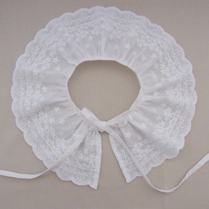 Può includere: Colletto in pizzo bianco con bordo a festoni e fiocco. Il colletto è realizzato in pizzo bianco delicato con un motivo floreale.