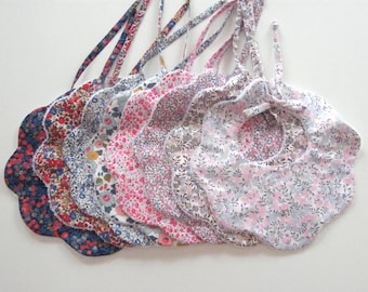 May include: A collection of floral print baby bibs in various colors, including red, blue, pink, and gray. Each bib has a scalloped edge and a tie closure at the neck.