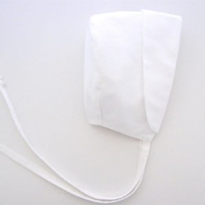 Puede incluir: Un gorro de bebé blanco hecho de tela suave. El gorro tiene una forma redondeada con un borde doblado y lazos largos y finos para asegurar debajo de la barbilla. El fondo es blanco liso.