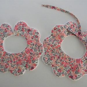 Peut inclure: Deux bavoirs pour bébé à motifs floraux, avec une bordure festonnée et une fermeture à nouer. Les bavoirs présentent un motif de petites fleurs roses et de feuilles vertes sur fond crème. Les bords sont garnis de tissu blanc.
