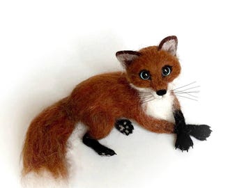 May include: A needle-felted fox figurine with rust-colored fur, a white chest, and black paws. The fox has large dark eyes, pointed ears with white inner details, and long thin whiskers. It is depicted in a resting position with its bushy tail extended.