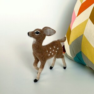 The picture shows a beautiful needle felted toy fawn made of wool, with a colorful pillow in the background.