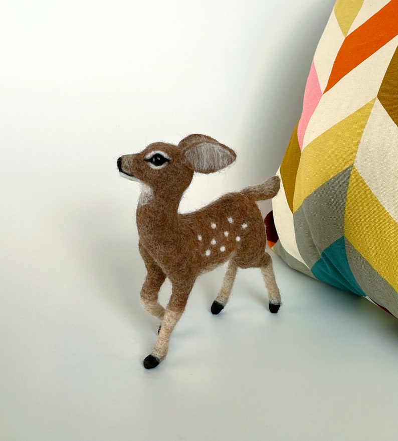 The picture shows a beautiful needle felted toy fawn made of wool, with a colorful pillow in the background.