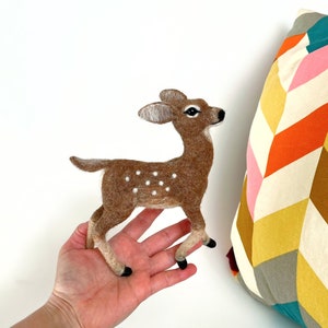 The picture shows a beautiful needle felted toy fawn made of wool standing on a palm, with a colorful pillow in the background.