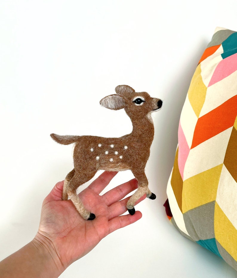 The picture shows a beautiful needle felted toy fawn made of wool standing on a palm, with a colorful pillow in the background.