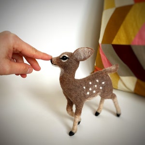 The image shows a beautiful needle felted toy deer made of wool reaching towards a hand, with a colorful pillow in the background.