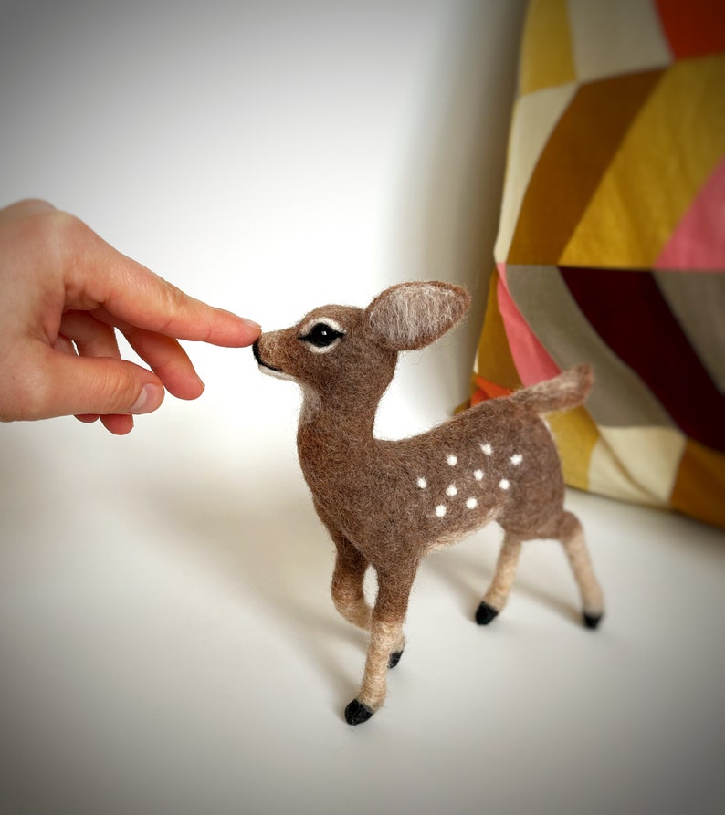The image shows a beautiful needle felted toy deer made of wool reaching towards a hand, with a colorful pillow in the background.