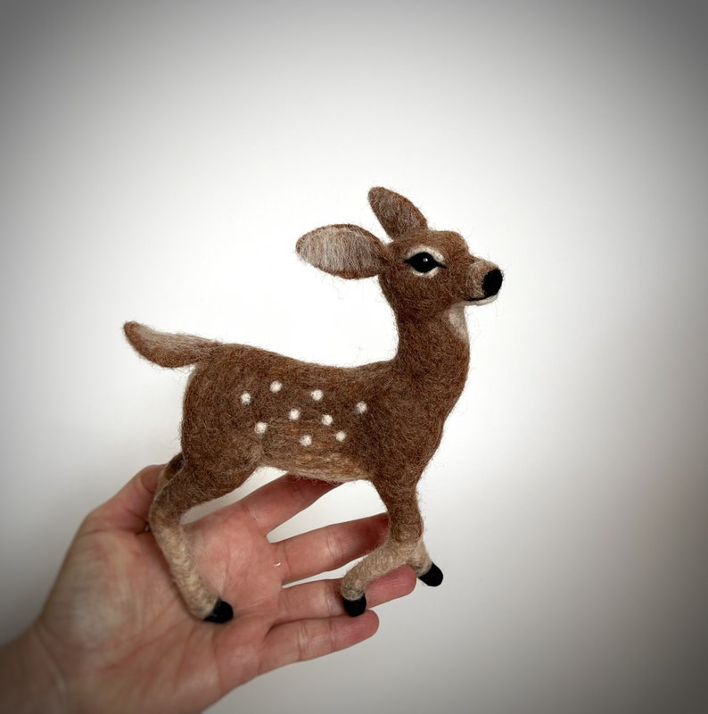 The picture shows a beautiful needle felted toy fawn made of wool standing on a palm