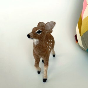 The picture shows a beautiful needle felted toy fawn made of wool, with a colorful pillow in the background.