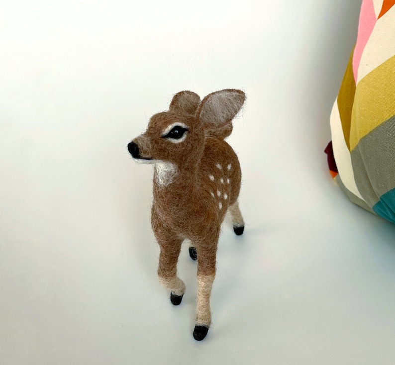 The picture shows a beautiful needle felted toy fawn made of wool, with a colorful pillow in the background.