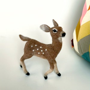 The picture shows a beautiful needle felted toy fawn made of wool, with a colorful pillow in the background.