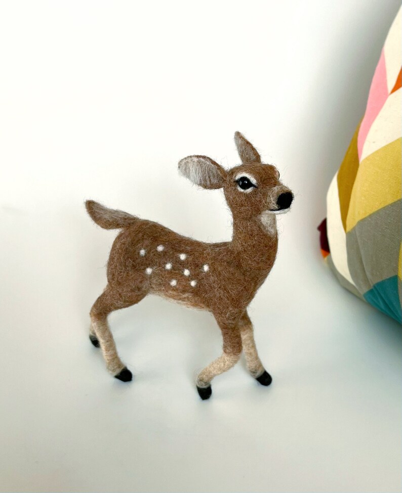 The picture shows a beautiful needle felted toy fawn made of wool, with a colorful pillow in the background.