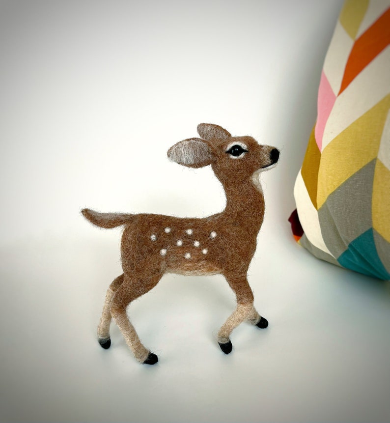 The picture shows a beautiful needle felted toy fawn made of wool, with a colorful pillow in the background.
