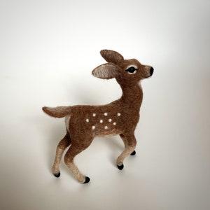 The picture shows a beautiful wool, needle felted toy fawn standing on a palm, the background is white.