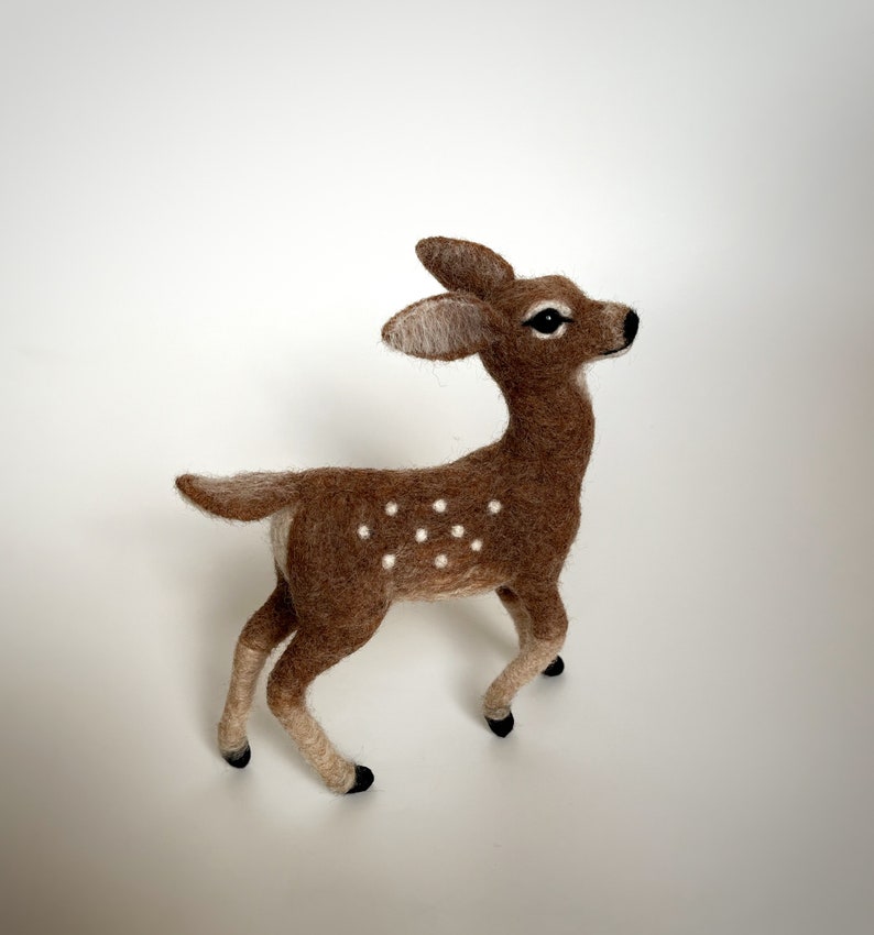 The picture shows a beautiful wool, needle felted toy fawn standing on a palm, the background is white.