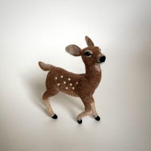 The picture shows a beautiful wool, needle felted toy fawn standing on a palm, the background is white.
