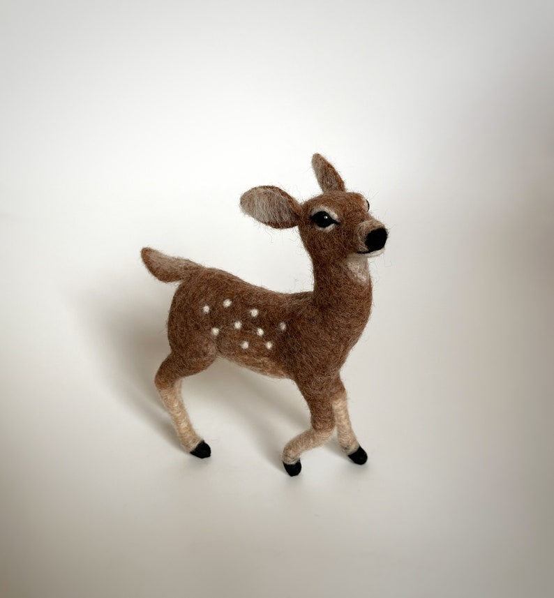 The picture shows a beautiful wool, needle felted toy fawn standing on a palm, the background is white.