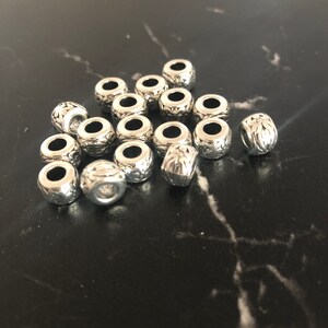 Op de afbeelding: Een groep zilveren metalen kralen met een decoratief ontwerp. De kralen zijn verspreid over een zwart oppervlak.