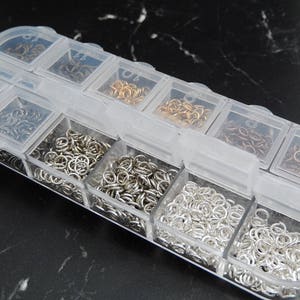 Op de afbeelding: Een transparante plastic opbergdoos met 10 vakken gevuld met verschillende maten sprongringen. De sprongringen zijn zilver, goud en brons.
