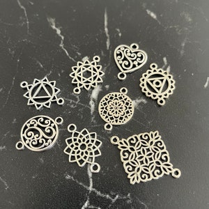Op de afbeelding: Een set van negen zilveren metalen bedels met ingewikkelde ontwerpen, waaronder een hart, een bloem, een vierkant en verschillende ronde ontwerpen. De bedels hebben elk drie gaten, wat suggereert dat ze bedoeld zijn voor het maken van sieraden.