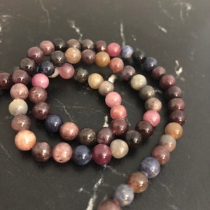 Könnte beinhalten: Eine Kette aus mehrfarbigen Edelsteinperlen, darunter Farbtöne von Rosa, Lila, Blau und Braun. Die Perlen sind rund und glatt und auf einer durchsichtigen Schnur aufgefädelt.