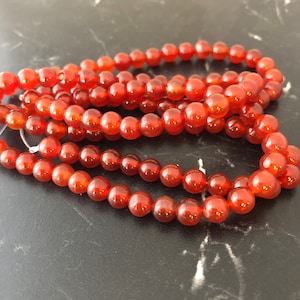 Peut inclure: Un brin de perles de cornaline, une variété de pierre précieuse, de couleur orange foncé. Les perles sont rondes et lisses, et elles sont disposées en un tas lâche.
