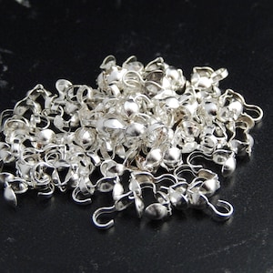 Könnte beinhalten: Ein Haufen silberfarbener Metallverschlüsse, jeder mit einer kleinen Schlaufe und einem abgerundeten Ende. Die Verschlüsse sind in einem zufälligen Muster angeordnet, wodurch ein strukturiertes und komplexes Design entsteht.