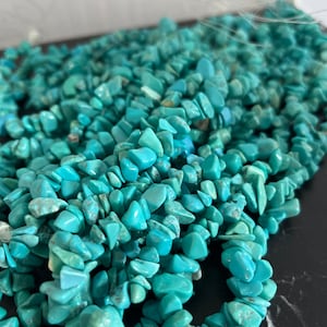 Pode incluir: Um fio de contas de turquesa em forma de lascas, com uma variedade de formas e tamanhos. As contas são de uma cor azul-verde vibrante e têm uma textura natural irregular.