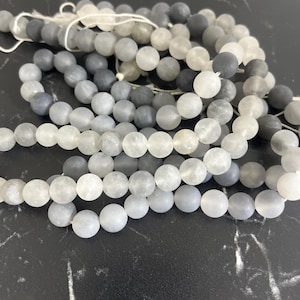 Könnte beinhalten: Eine Strähne aus grauen und weißen, mattierten Glasperlen. Die Perlen sind rund und haben eine matte Oberfläche.