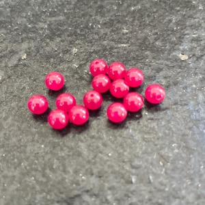 Può includere: Un insieme di piccole perline rotonde rosa acceso sparse su una superficie scura e strutturata. Ogni perlina ha una finitura lucida e un piccolo foro, suggerendo che siano per infilare o creare. Le perline sono di dimensioni uniformi.