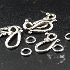 Op de afbeelding: Vijf zilverkleurige metalen sluitingen met sierlijke ontwerpen. De sluitingen hebben de vorm van een lus met een decoratieve swirl. Er zijn ook vier zilverkleurige metalen springringen.