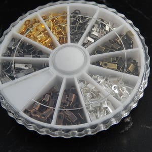 Op de afbeelding: Een doorzichtige, gesegmenteerde container gevuld met diverse metalen krimpeindstukken in goud-, zilver- en bronstinten. De eindstukken zijn klein, rechthoekig en worden gebruikt voor het maken van sieraden. De container staat op een zwart oppervlak.