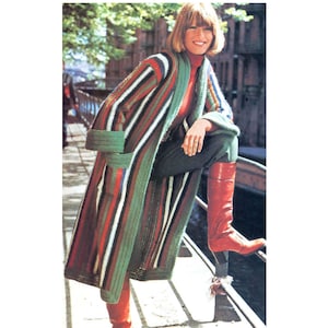 May include: A long, striped, green, orange, and white cardigan with a shawl collar. The cardigan is worn by a person with blonde hair and is standing on a wooden walkway.