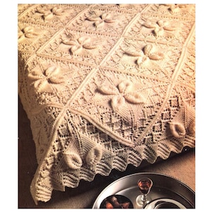 May include: A crocheted throw blanket with a delicate floral pattern in a cream color. The blanket is draped over a bed and has a scalloped edge.