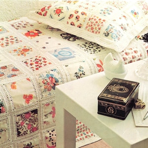 K&ouml;nnte beinhalten: Ein Patchwork-Quilt und Kissen-Set mit bunten Blumenmustern. Der Quilt hat quadratische Flicken mit verschiedenen Blumendesigns, die von wei&szlig;er H&auml;kelarbeit umrandet sind. Ein kleiner wei&szlig;er Tisch mit einer dekorativen Box, W&uuml;rfeln, einem Stift und einem Notizblock.