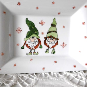 May include: White rectangular ceramic plate featuring two cartoon gnomes in green hats and red coats, with red snowflake motifs. A whimsical design for festive holiday decorations.