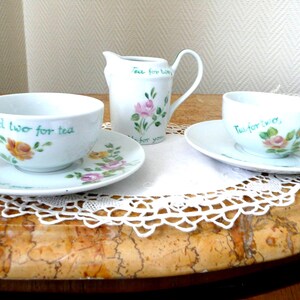 Peut inclure: Service à thé en porcelaine blanche avec des motifs floraux et l'inscription "Tea for two". L'ensemble comprend deux tasses et soucoupes, et un pot à crème. Le motif floral présente des roses roses et jaunes.