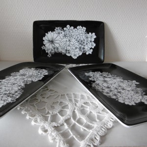 Peut inclure: Trois plateaux de service rectangulaires noirs avec des motifs floraux blancs. Les plateaux sont disposés sur une surface blanche avec une dentelle blanche en crochet.