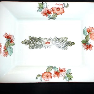 Peut inclure: Assiette rectangulaire en céramique blanche avec un bord rouge et des motifs floraux et celtiques. Les coins présentent des grappes de fleurs orange et roses avec des feuilles vertes. Un motif central de nœud celtique est mis en valeur par un ovale rouge.